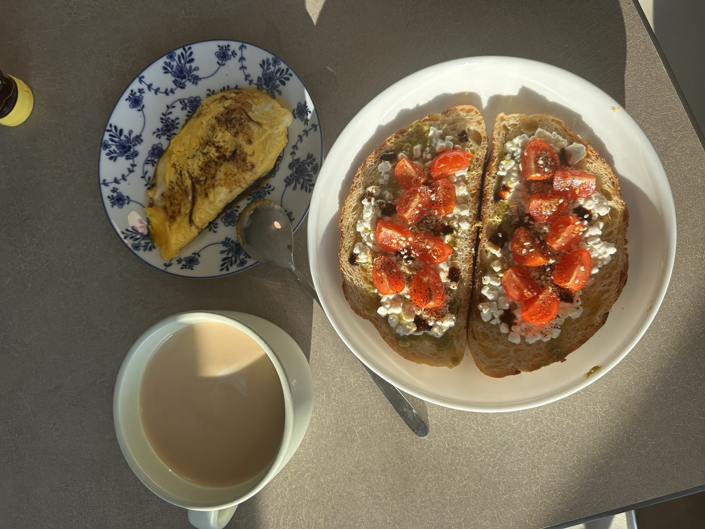 Breakfast in morning light: sourdough toast topped with cottage cheese, cherry tomatoes and balsamic, a folded omelette, and a mug of coffee