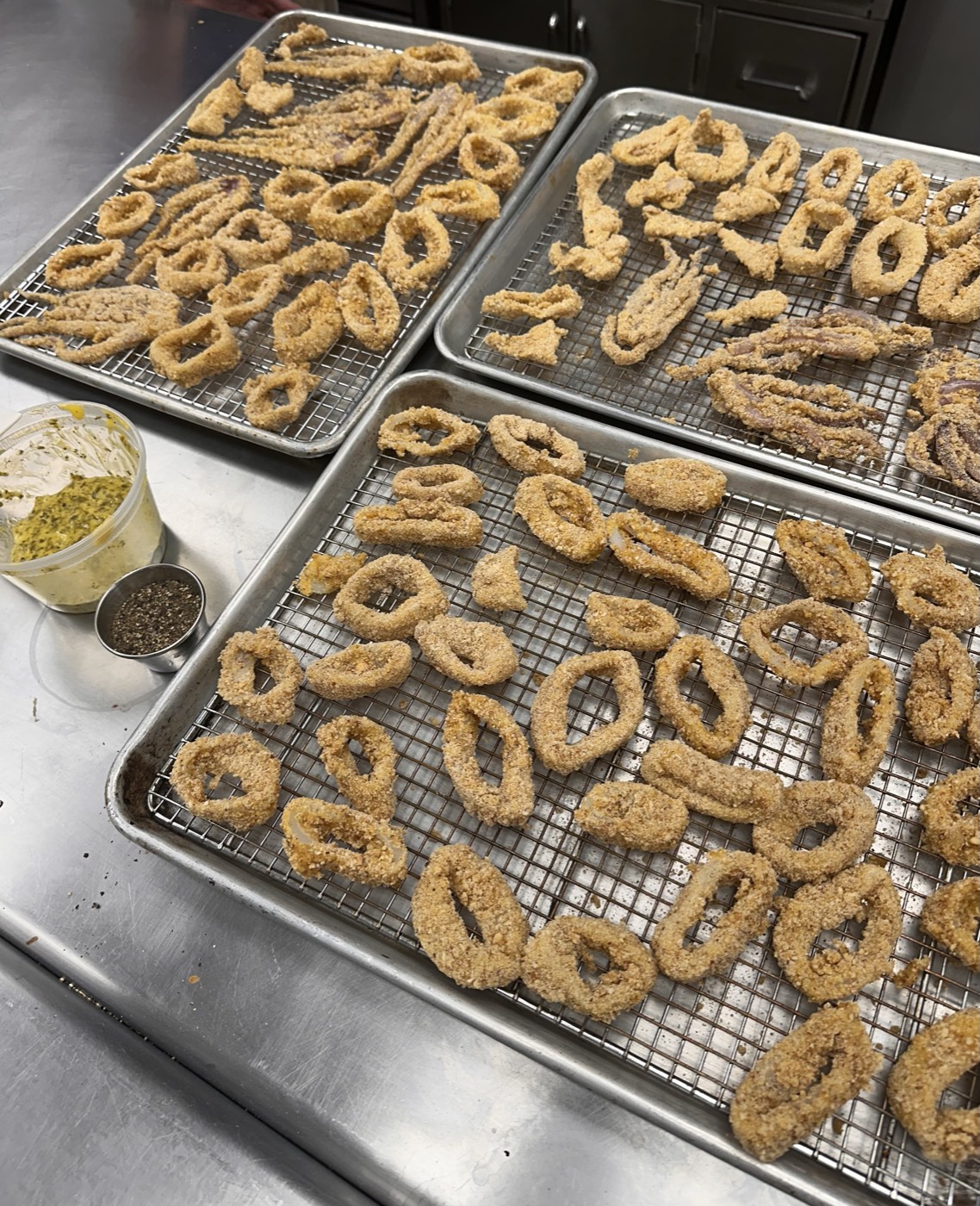 Three sheet pans of breaded calamari rings and tentacles resting on wire racks in a kitchen, ready for the fryer