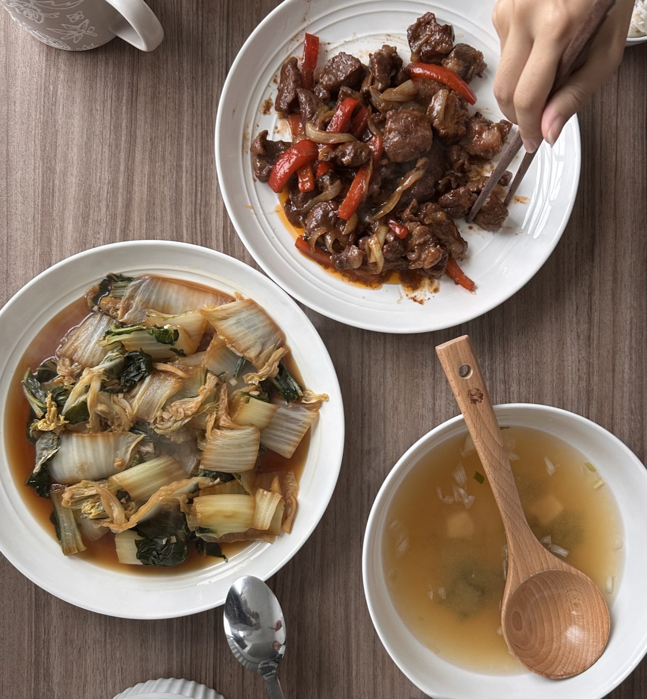Dinner spread on a wooden table: beef and red pepper stir-fry, braised napa cabbage in broth, and a bowl of tofu and scallion soup